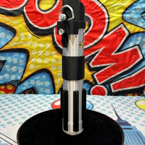 Może przedstawiać: Srebrno-czarna replika miecza świetlnego z czerwonym ostrzem, prezentowana na czarnym, okrągłym stojaku. Rękojeść posiada szczeg&oacute;łowe przyciski i pierścień. Tło to kolorowy wz&oacute;r pop-art.