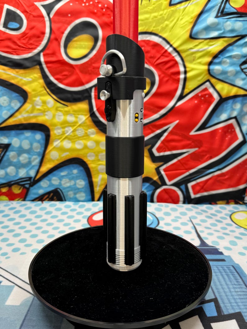 Może przedstawiać: Srebrno-czarna replika miecza świetlnego z czerwonym ostrzem, prezentowana na czarnym, okrągłym stojaku. Rękojeść posiada szczeg&oacute;łowe przyciski i pierścień. Tło to kolorowy wz&oacute;r pop-art.