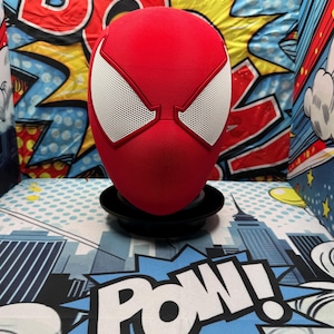 May include: A red full-head mask featuring large, white, mesh-covered eye lenses. The mask has a textured, fabric-like surface and is mounted on a black circular stand. The design is inspired by a superhero character, showcasing a sleek, rounded shape.