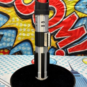 Może przedstawiać: Replika miecza świetlnego z czerwonym ostrzem, srebrnym i czarnym uchwytem, na czarnej okrągłej podstawie. Rękojeść ma szczeg&oacute;łowe srebrne i czarne elementy. Tło to kolorowy wz&oacute;r pop-art.