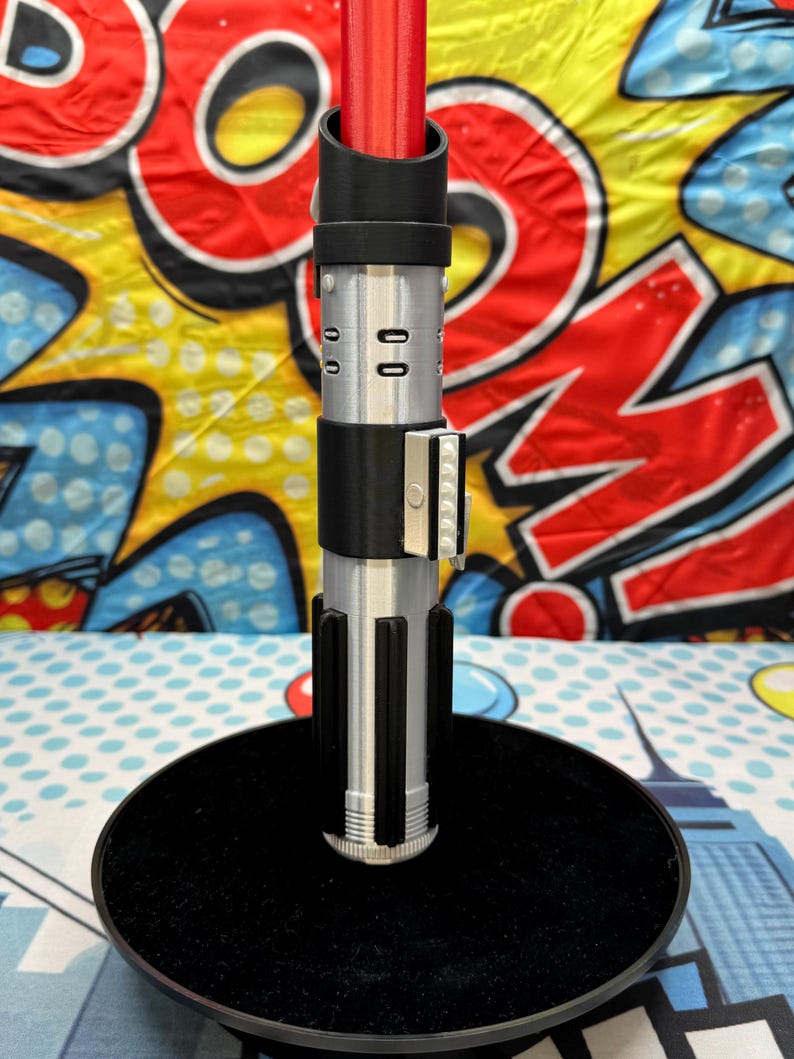 Może przedstawiać: Replika miecza świetlnego z czerwonym ostrzem, srebrnym i czarnym uchwytem, na czarnej okrągłej podstawie. Rękojeść ma szczeg&oacute;łowe srebrne i czarne elementy. Tło to kolorowy wz&oacute;r pop-art.