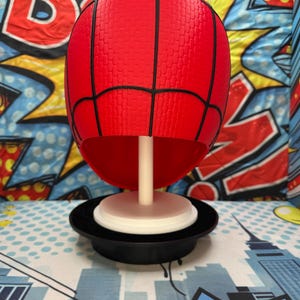 May include: A red Spider-Man mask replica with black webbing lines, mounted on a white stand and a black base. The mask has a textured surface and is displayed against a colorful comic book-themed backdrop.