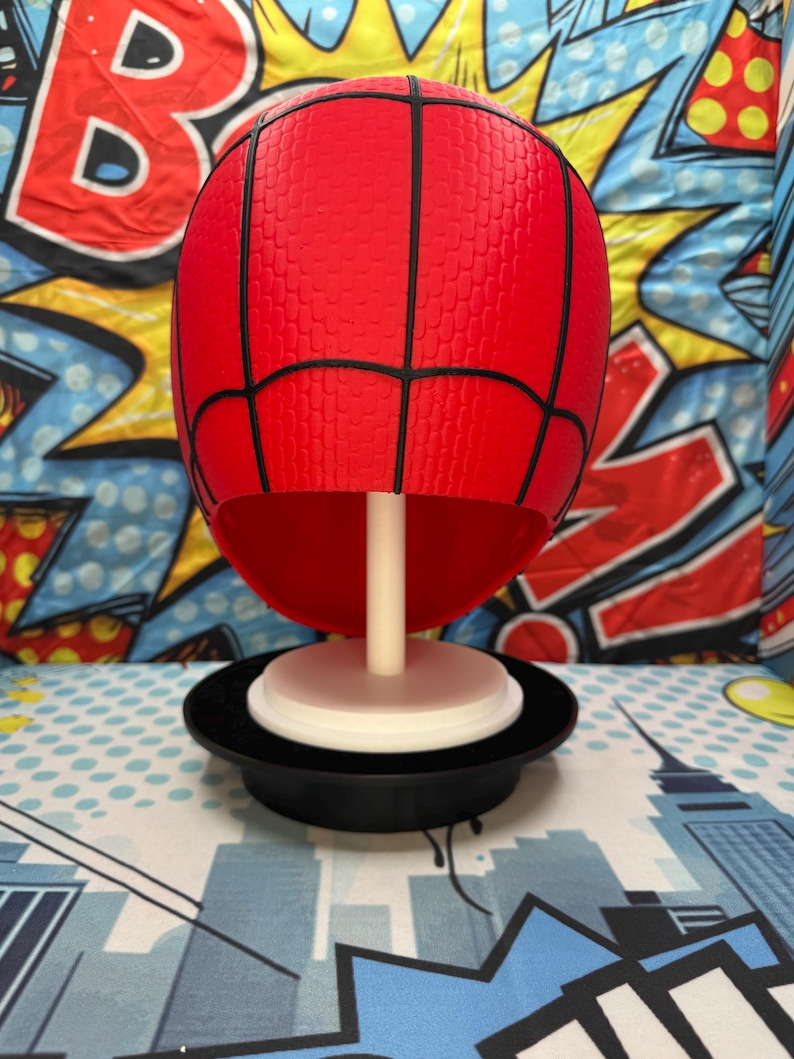 May include: A red Spider-Man mask replica with black webbing lines, mounted on a white stand and a black base. The mask has a textured surface and is displayed against a colorful comic book-themed backdrop.
