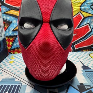 Könnte beinhalten: Eine rote Deadpool-Maske mit schwarzen Augenabdeckungen und weißen Netzeinsätzen. Die Maske hat eine strukturierte Oberfläche und wird auf einem schwarzen Ständer präsentiert. Der Hintergrund zeigt farbenfrohe Grafiken im Comic-Stil.