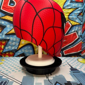 May include: A red Spider-Man mask with black webbing details, displayed on a white stand with a black base. The mask is a vibrant red with a textured surface. The background features a comic book-style design.