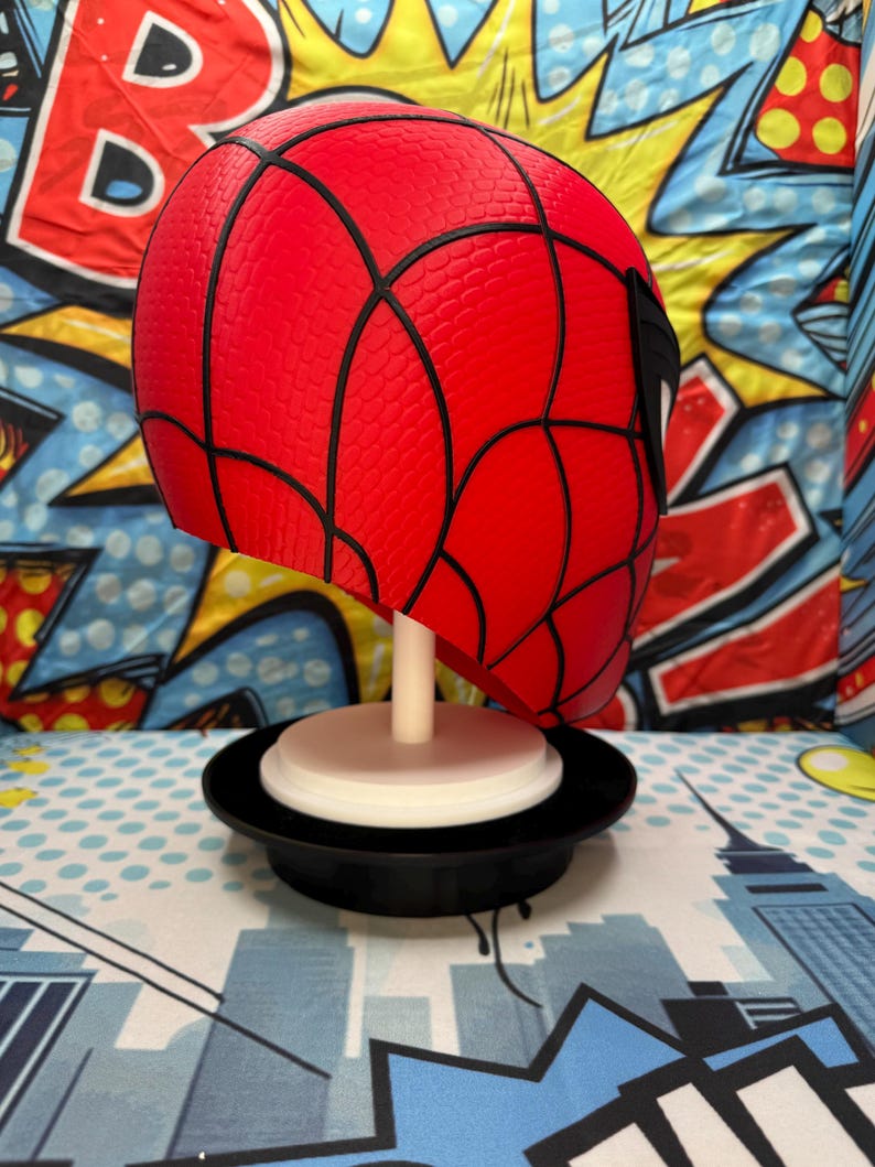 May include: A red Spider-Man mask with black webbing details, displayed on a white stand with a black base. The mask is a vibrant red with a textured surface. The background features a comic book-style design.