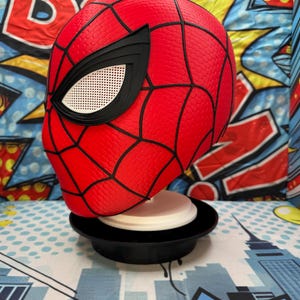 May include: A red Spider-Man mask with black webbing and eye details. The mask is displayed on a black stand with a white base, set against a colorful comic book-style backdrop.