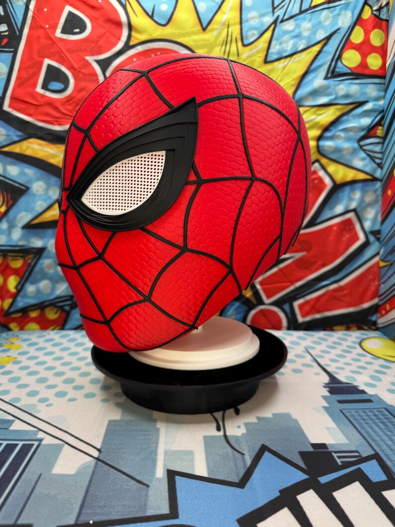 May include: A red Spider-Man mask with black webbing and eye details. The mask is displayed on a black stand with a white base, set against a colorful comic book-style backdrop.