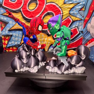 May include: A 3D-printed Spider-Man figure in red, blue, and white, facing off against a green and purple Green Goblin figure. Both figures are on a black platform with a comic book-style backdrop. The figures are posed in action.