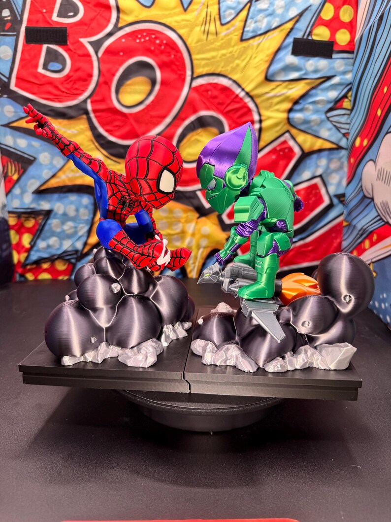May include: A 3D-printed Spider-Man figure in red, blue, and white, facing off against a green and purple Green Goblin figure. Both figures are on a black platform with a comic book-style backdrop. The figures are posed in action.