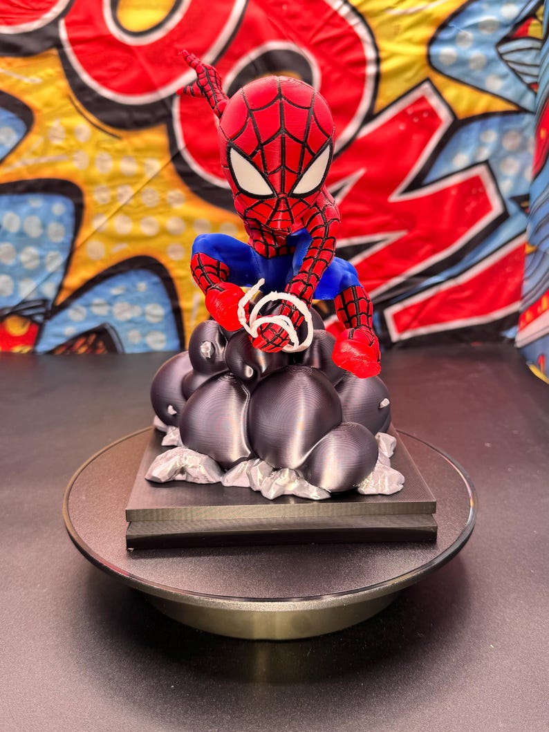 May include: A red, blue, and white Spider-Man figurine, poised as if shooting a web. The figurine is atop a black base with dark gray spheres and silver accents. The background features a colorful comic book-style design.