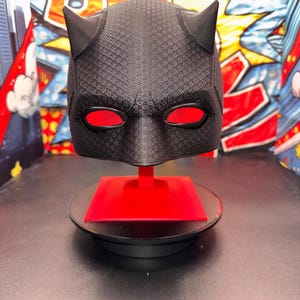 Puede incluir: Un casco negro texturizado con dos cuernos prominentes en la parte superior y lentes oculares de malla roja vibrante. El casco está montado sobre un soporte cuadrado rojo, colocado sobre una base circular negra. La superficie tiene un patrón de rejilla.