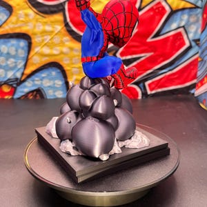 May include: A 3D-printed Spider-Man figurine in a dynamic pose, featuring a red and blue suit. The figure is perched atop a cluster of dark gray spheres and a square base, set against a colorful comic-style backdrop.
