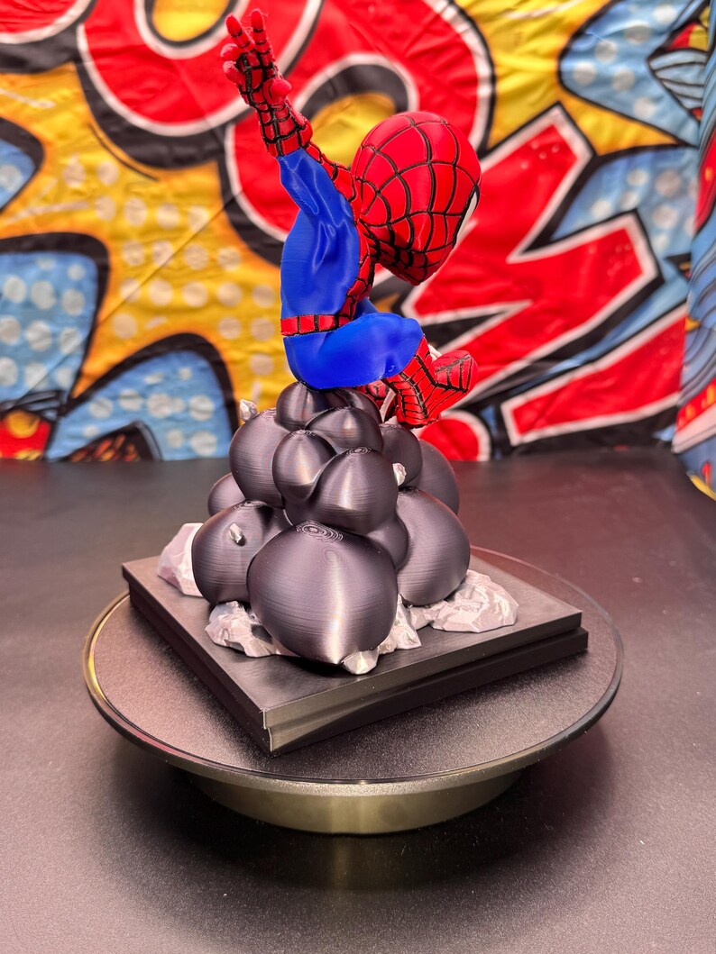 May include: A 3D-printed Spider-Man figurine in a dynamic pose, featuring a red and blue suit. The figure is perched atop a cluster of dark gray spheres and a square base, set against a colorful comic-style backdrop.