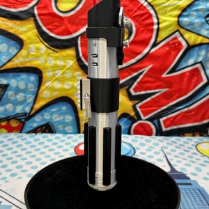 Może przedstawiać: Srebrna i czarna replika miecza świetlnego z czerwonym ostrzem, prezentowana na czarnym, okrągłym stojaku. Rękojeść posiada szczeg&oacute;łowe srebrne i czarne elementy, w tym panel sterowania i osłonę emiter&oacute;w. Tło to kolorowy design pop-art.