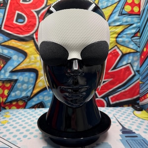 Puede incluir: Un busto de maniqu&iacute; negro exhibe una m&aacute;scara de ojos blanca y negra. La m&aacute;scara presenta una secci&oacute;n superior blanca texturizada y dos cubiertas oculares de malla negra. El maniqu&iacute; est&aacute; sobre una base negra, sobre un fondo de arte pop colorido.