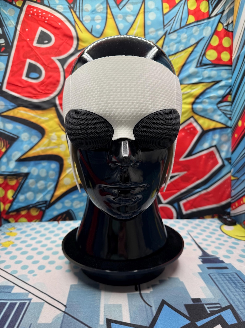 Puede incluir: Un busto de maniqu&iacute; negro exhibe una m&aacute;scara de ojos blanca y negra. La m&aacute;scara presenta una secci&oacute;n superior blanca texturizada y dos cubiertas oculares de malla negra. El maniqu&iacute; est&aacute; sobre una base negra, sobre un fondo de arte pop colorido.