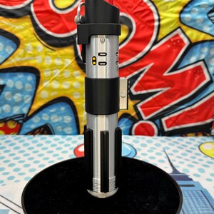 Może przedstawiać: Srebrno-czarna replika miecza świetlnego z czerwonym ostrzem, prezentowana na czarnym stojaku. Rękojeść posiada szczeg&oacute;łowe elementy i elegancki design. Tło jest w kolorowym stylu pop-art.