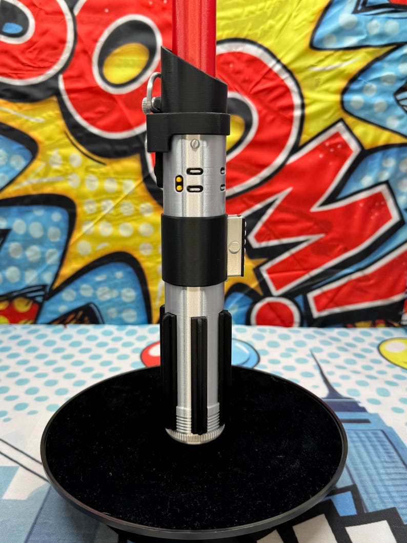 Może przedstawiać: Srebrno-czarna replika miecza świetlnego z czerwonym ostrzem, prezentowana na czarnym stojaku. Rękojeść posiada szczeg&oacute;łowe elementy i elegancki design. Tło jest w kolorowym stylu pop-art.