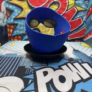 Puede incluir: Un accesorio azul en forma de casco con cubiertas oculares de malla, apoyado en un platillo negro. El fondo presenta un dise&ntilde;o de estilo c&oacute;mic con la palabra "POW" en blanco y un tel&oacute;n de fondo con la palabra "BOOM".