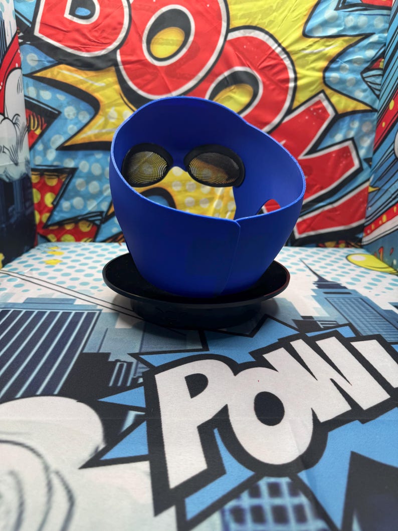 Puede incluir: Un accesorio azul en forma de casco con cubiertas oculares de malla, apoyado en un platillo negro. El fondo presenta un dise&ntilde;o de estilo c&oacute;mic con la palabra "POW" en blanco y un tel&oacute;n de fondo con la palabra "BOOM".