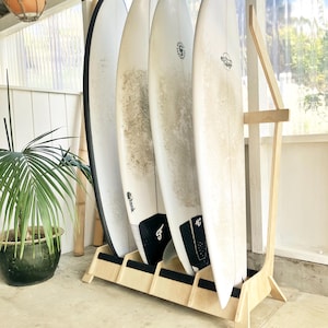 Könnte beinhalten: Ein Surfboard-Rack aus Holz mit vier Surfboards, die dagegen gelehnt sind. Das Rack ist aus hellem Holz und hat ein schlichtes Design. Die Surfboards sind alle weiß und haben schwarze Finnen.