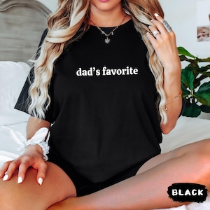 May include: Black t-shirt with the text "dad's favorite" in white. The shirt is a crew neck style and appears to be made of a soft material. The word "BLACK" is in a black and white rectangle at the bottom of the image.