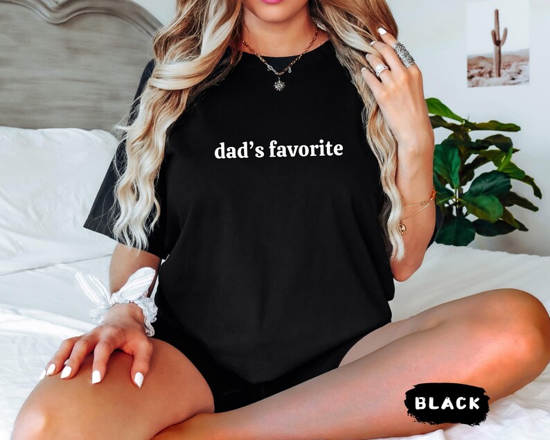 May include: Black t-shirt with the text "dad's favorite" in white. The shirt is a crew neck style and appears to be made of a soft material. The word "BLACK" is in a black and white rectangle at the bottom of the image.