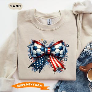 May include: Sand-colored sweatshirt featuring a soccer ball bow design with red, white, and blue accents. The bow is surrounded by soccer balls and a patriotic color scheme. The text "SHIPS NEXT DAY!" is also visible.