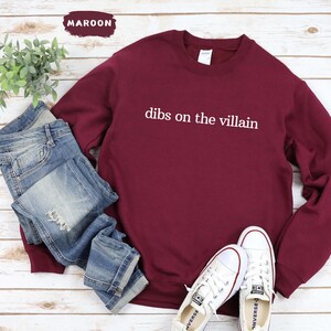 May include: A maroon sweatshirt with the text "dibs on the villain" in white. The sweatshirt is styled with blue jeans and white trainers. A small plant is visible in the background.