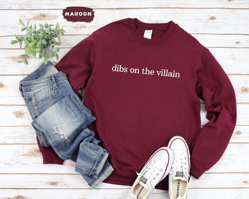 May include: A maroon sweatshirt with the text "dibs on the villain" in white. The sweatshirt is styled with blue jeans and white trainers. A small plant is visible in the background.