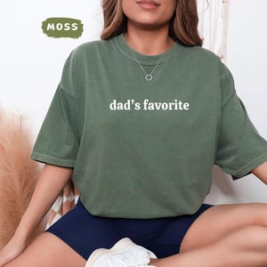 May include: Moss green t-shirt with the text "dad's favorite" in white. The shirt is a relaxed fit and has short sleeves. The word "MOSS" is in a green rectangle in the upper left corner.