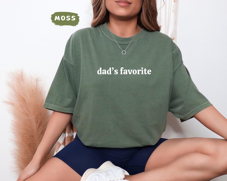 May include: Moss green t-shirt with the text "dad's favorite" in white. The shirt is a relaxed fit and has short sleeves. The word "MOSS" is in a green rectangle in the upper left corner.