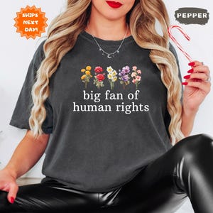 May include: Dark gray t-shirt with the text "big fan of human rights" and a floral design. The flowers include yellow, red, white, and purple blooms. The shirt is a comfortable, casual style.