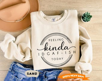 May include: A sand-colored crewneck sweatshirt featuring a black circular graphic with the text "FEELING kinda IDGAF-ISH TODAY" printed in the center. The sweatshirt has long sleeves and a ribbed collar, cuffs, and hem, presented in a neutral tone.