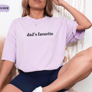 May include: A light purple t-shirt with the text "dad's favorite" in black. The shirt is a relaxed fit and is paired with navy blue shorts. The word "ORCHID" is in a purple box in the upper left corner.