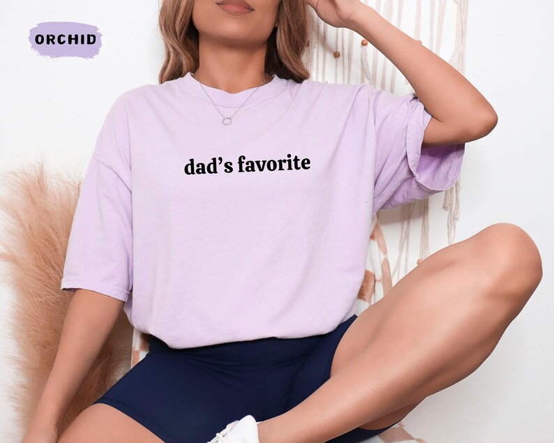 May include: A light purple t-shirt with the text "dad's favorite" in black. The shirt is a relaxed fit and is paired with navy blue shorts. The word "ORCHID" is in a purple box in the upper left corner.