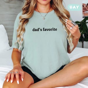 May include: A light green t-shirt with the text "dad's favorite" in black. The shirt is a crew neck style and appears to be made of a soft material. The word "BAY" is in a light green box in the upper right corner.