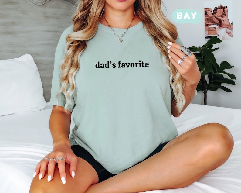 May include: A light green t-shirt with the text "dad's favorite" in black. The shirt is a crew neck style and appears to be made of a soft material. The word "BAY" is in a light green box in the upper right corner.