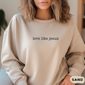 May include: A sand-colored sweatshirt with the text "love like jesus" printed in black. The sweatshirt has a crew neck and long sleeves. The word "SAND" is printed in a white box in the lower right corner.