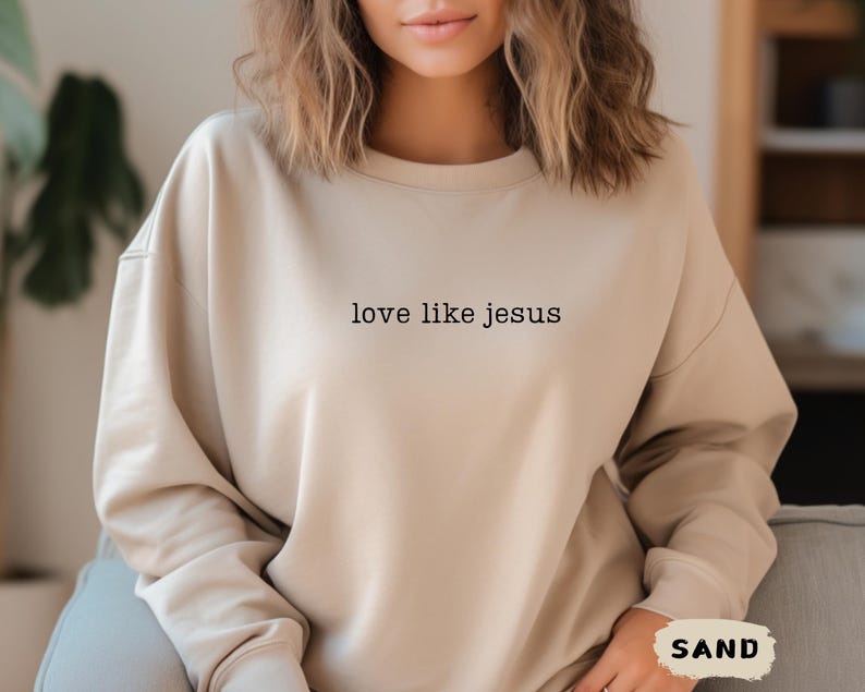 May include: A sand-colored sweatshirt with the text "love like jesus" printed in black. The sweatshirt has a crew neck and long sleeves. The word "SAND" is printed in a white box in the lower right corner.