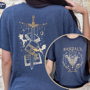 May include: Heather navy blue t-shirt with a gold design of a sword, books, and bats on the back. A circular inset shows a matching shirt with the text "BASGIATH WAR COLLEGE" and a dragon design. The shirt is a fantasy theme.