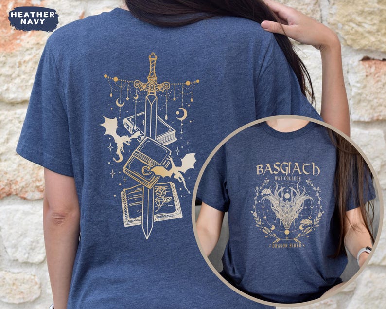 May include: Heather navy blue t-shirt with a gold design of a sword, books, and bats on the back. A circular inset shows a matching shirt with the text "BASGIATH WAR COLLEGE" and a dragon design. The shirt is a fantasy theme.
