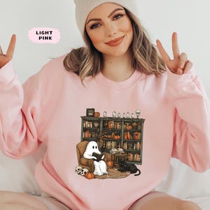 May include: Light pink sweatshirt featuring a Halloween-themed graphic. The graphic depicts a ghost reading in a cozy armchair in front of a bookshelf, with pumpkins, candles, and a black cat. The text "LIGHT PINK" is in a pink box.