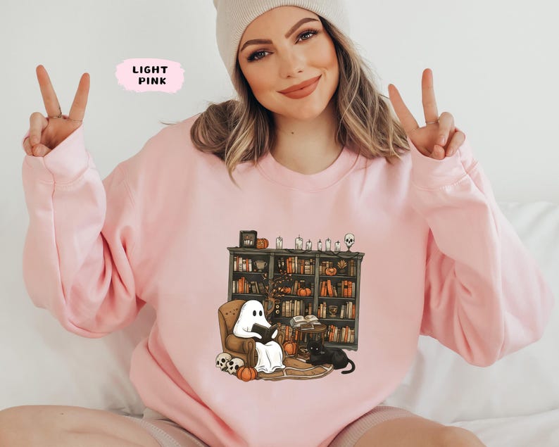 May include: Light pink sweatshirt featuring a Halloween-themed graphic. The graphic depicts a ghost reading in a cozy armchair in front of a bookshelf, with pumpkins, candles, and a black cat. The text "LIGHT PINK" is in a pink box.