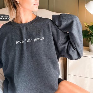 May include: Heather dark grey sweatshirt with the text "love like jesus" in white. The sweatshirt is a crew neck style and has long sleeves. The image shows the sweatshirt being worn.