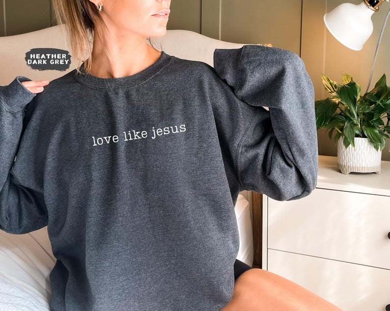 May include: Heather dark grey sweatshirt with the text "love like jesus" in white. The sweatshirt is a crew neck style and has long sleeves. The image shows the sweatshirt being worn.