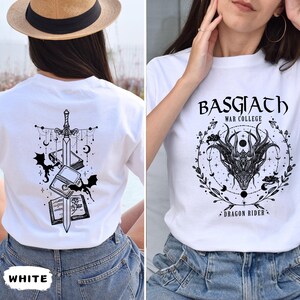 May include: White t-shirts with graphic designs. One shirt features a sword, books, bats, and celestial elements. The other shirt displays the text "BASGIATH WAR COLLEGE DRAGON RIDER" with a dragon head and floral accents.