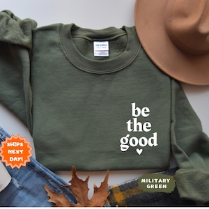May include: A green sweatshirt with the text "be the good" in white lettering and a small heart symbol. The sweatshirt is on a white background with a brown hat and blue jeans.