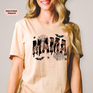 May include: Heather peach-colored t-shirt with the word "MAMA" in a dripping, leopard print design. The shirt features spiderwebs, spiders, and bats, creating a Halloween theme. The shirt is a short-sleeved crew neck.